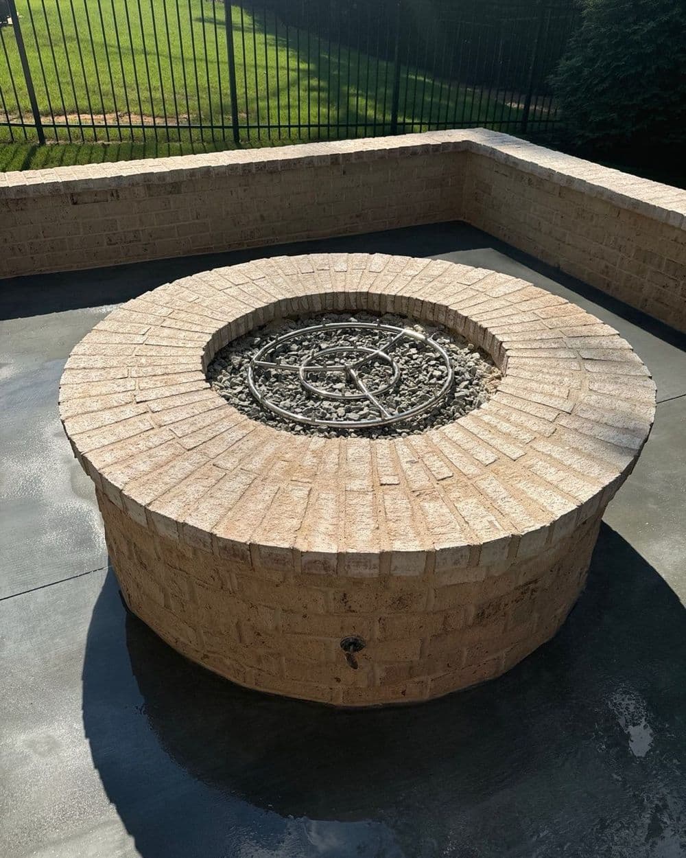 Stone fire pit with a decorative metal grate surrounded by a patio in a sunny backyard.